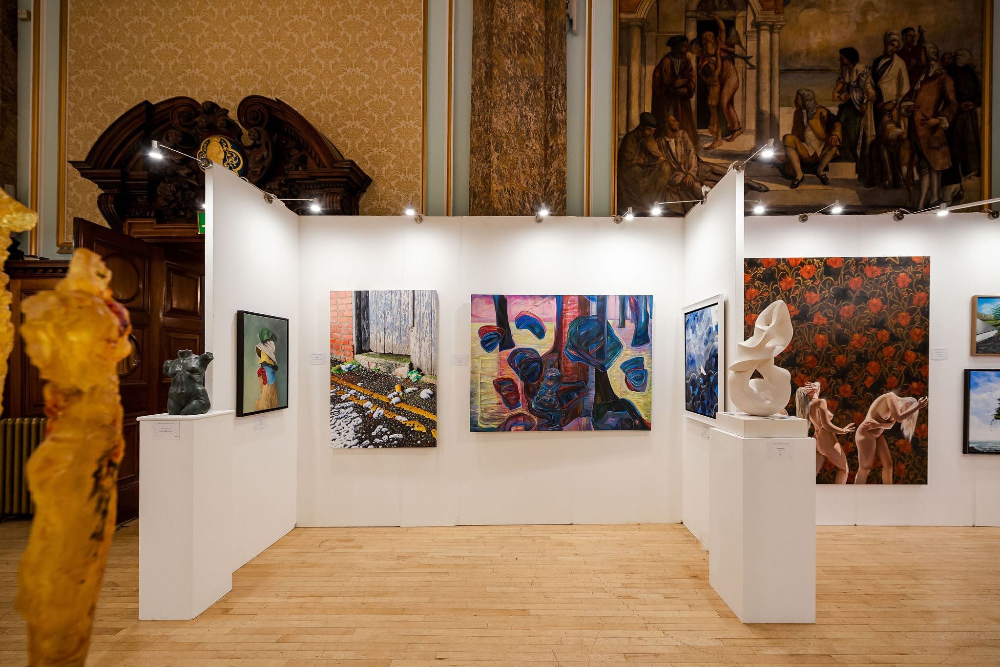 A corner of the exhibition: paintings and sculpture on white walls beneath a mural in the main hall.