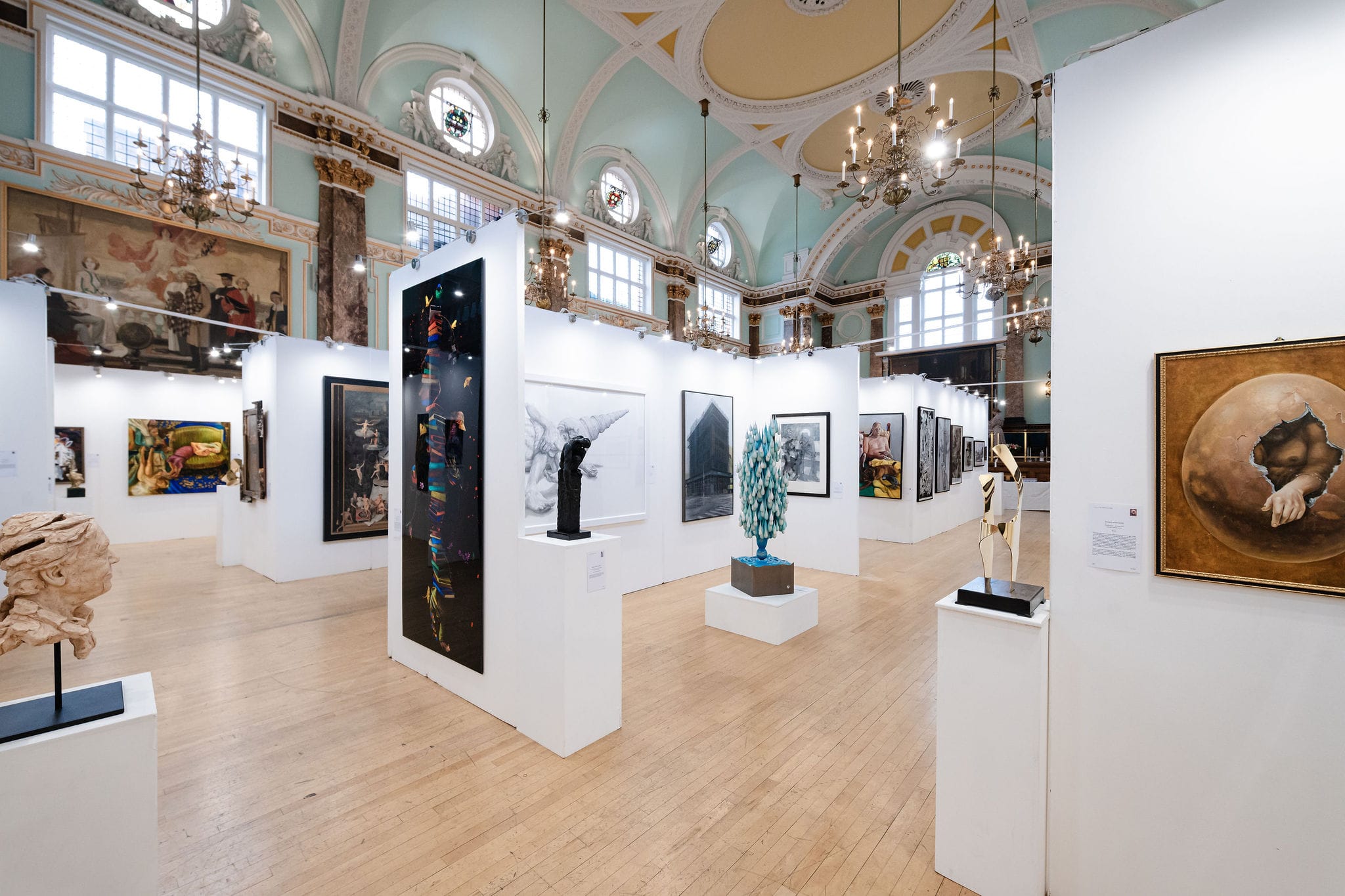 The main hall hung for the 2025 edition: sculpture on plinths between white walls of paintings, under the chandeliers and the painted ceiling.