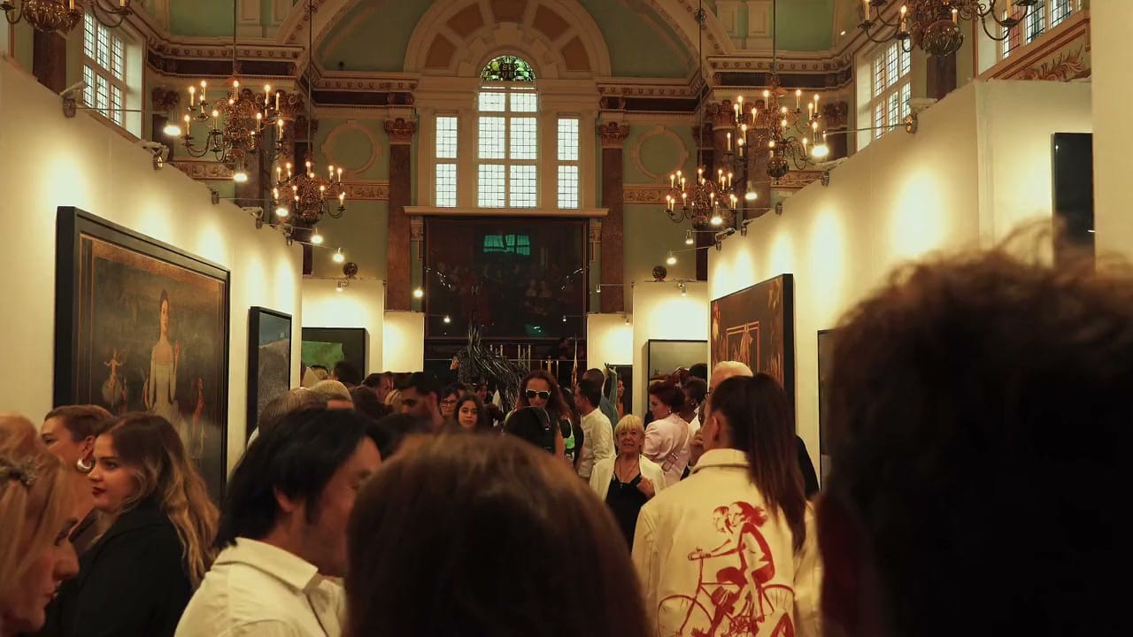The main hall at the Seventh Edition's opening in 2025, packed with visitors beneath the chandeliers and the arched window, paintings hung along both walls.