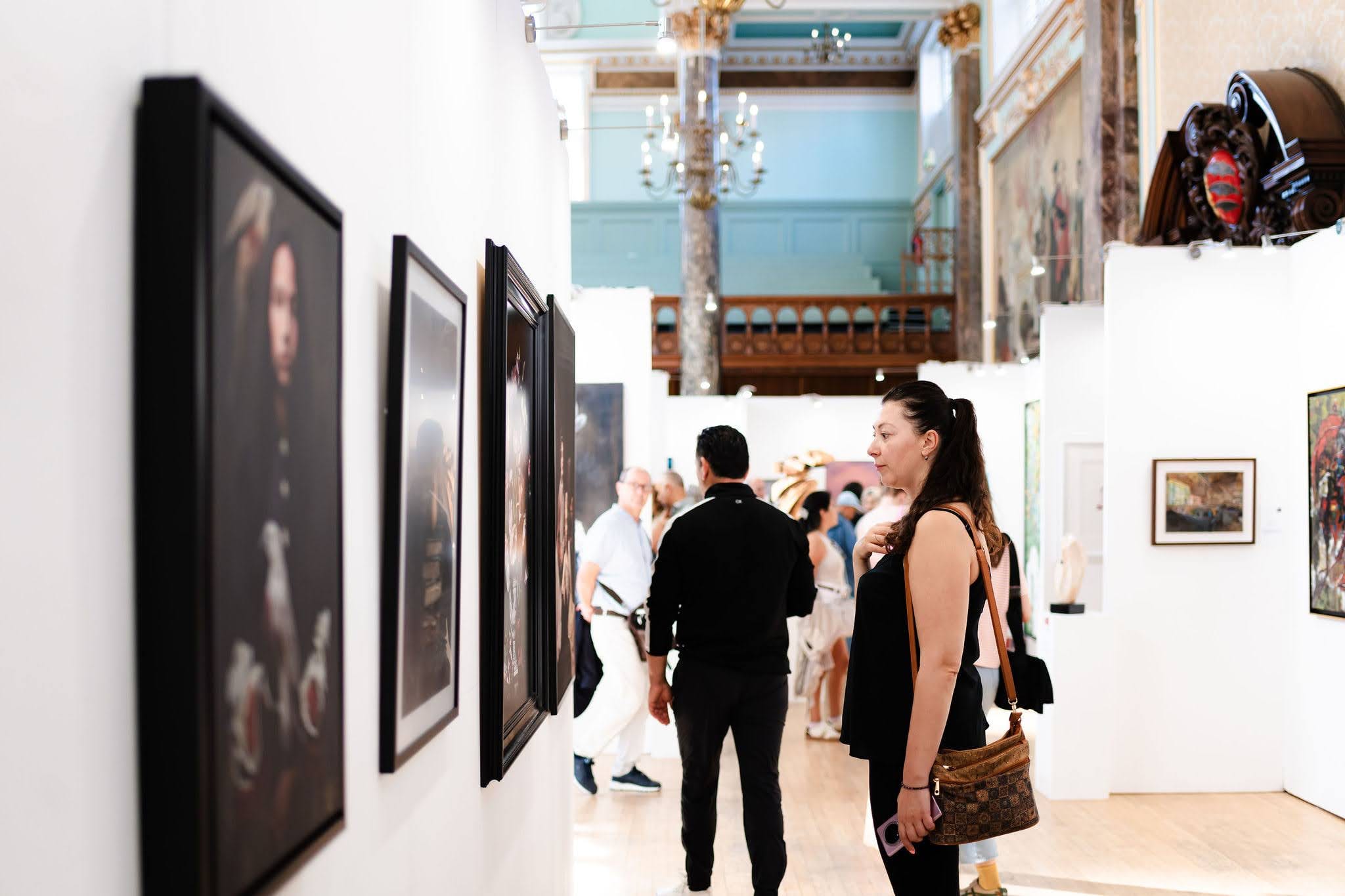 A visitor studying a wall of framed paintings, the hang and the balcony of the hall behind her.