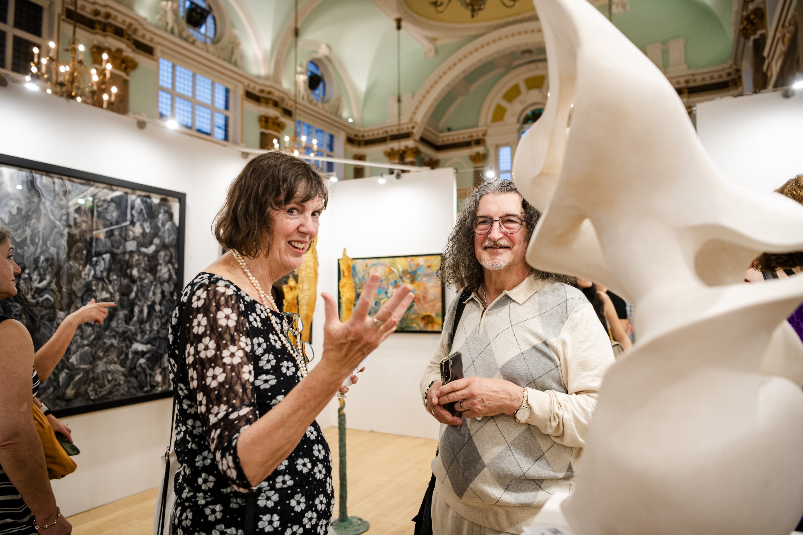 Two visitors deep in conversation at the opening, a white marble sculpture curving into the foreground.
